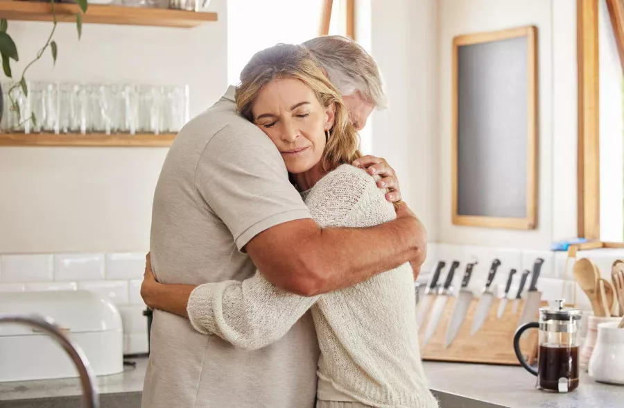 Grieving elderly couple hugging in kitchen to support one another.
